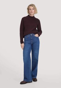Maglione lavorato a maglia bordeaux con collo alto, abbinato a jeans blu a gamba larga. Il modello indossa calzature marroni, in piedi davanti a uno sfondo chiaro.
