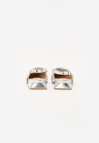 Pantofi plata metalici argintii cu un finisaj texturat, având un toc mic și o curea reglabilă la gleznă cu accente decorative.