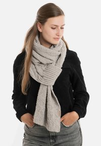 Lichtgrijze gebreide sjaal met een gestructureerd oppervlak en ribbelzoom, losjes om de nek gedragen, gecombineerd met een zwart topje en denim.