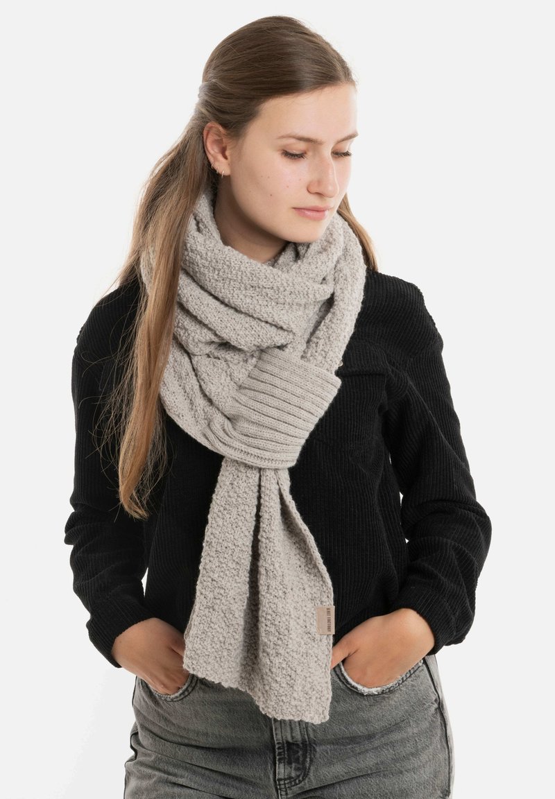 Lichtgrijze gebreide sjaal met een gestructureerd oppervlak en ribbelzoom, losjes om de nek gedragen, gecombineerd met een zwart topje en denim.