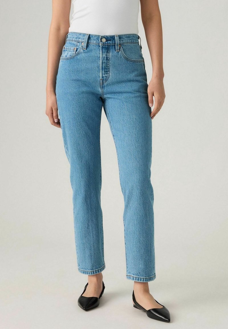 Personne portant un jean bleu clair taille haute à jambes droites, un haut blanc sans manches et des chaussures plates noires à bouts pointus, debout sur un fond uni.