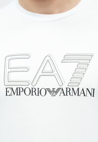 Valge lühikeste varrukatega T-särk musta graafilise disainiga, mis sisaldab teksti "EA7" ja "EMPORIO ARMANI" erineva joonte paksuse ja stiilidega.