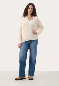 Cream knitted sweater with a V-neck and long sleeves, paired with blue wide-leg jeans and brown strappy sandals on a neutral background.