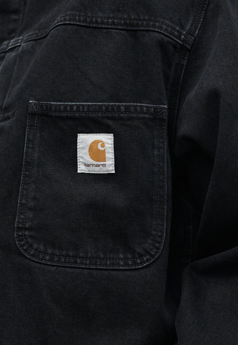 Tissu noir avec une poche carrée cousue affichant une étiquette blanche portant un logo doré partiellement incurvé et le texte "carhartt".