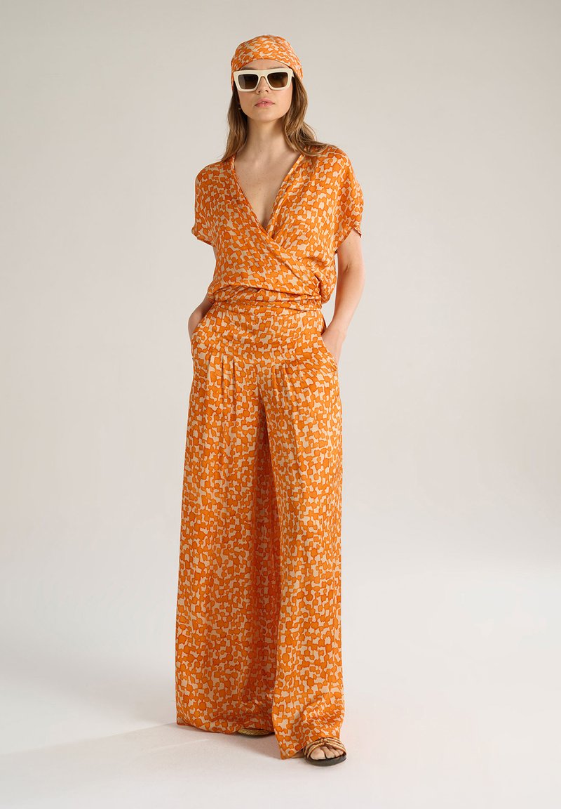 Top avvolgente arancione a fantasia con pantaloni a gamba larga, realizzato in un tessuto morbido, con scollatura a V profonda e tasche laterali. Indossato con occhiali da sole e un foulard.