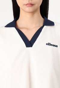 Vit pikétröja i frotté med marinblå krage och broderad "ellesse"-logotyp i marinblått. Har en distinkt V-halsdesign.