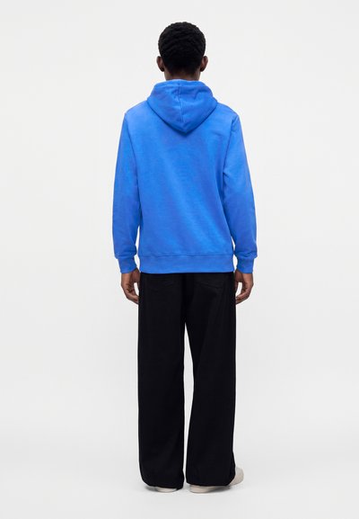 Homme vu de dos portant un sweat à capuche bleu vif, un pantalon noir large et des chaussures claires sur fond blanc uni.