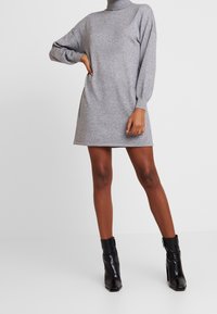 Robe en tricot gris avec un col montant, des manches longues et une coupe décontractée, associée à des bottines noires à la cheville. Texture lisse, longueur mi-cuisse.