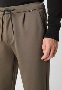 Smeđe hlače napravljene od teksturiranog materijala, s pojasom na vezanje, prednjim naborima i oznakom Calvin Klein na pojasu.