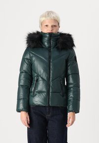 Green padded jacket with a high neck and black faux fur hood. Features diagonal chevron stitching and two zippered side pockets.