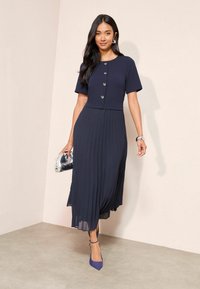 Tengerészkék pliszírozott ruha, testhezálló fazonnal és rövid ujjakkal, szív alakú gombokkal díszítve; ezüst clutch táskával és kék magassarkúval párosítva.