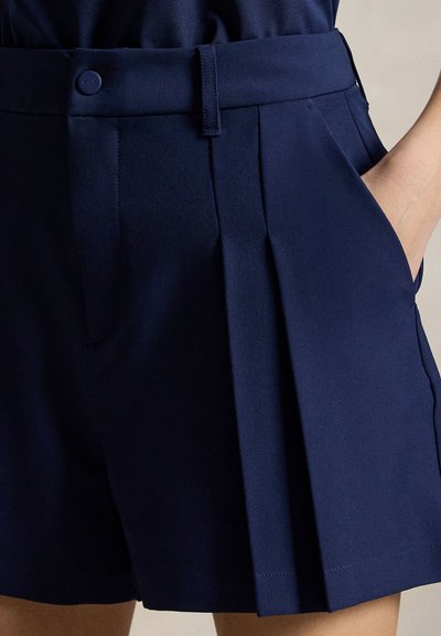Tmavomodré plisované šortky s bočnými vreckami, pútkami na opasok a zapínaním na gombík, nosené so zastrčeným tmavomodrým topom na mieru.