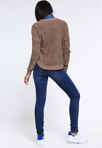Suéter de punto marrón con un cuello amplio y puños acanalados, combinado con jeans skinny azul oscuro y zapatillas blancas con detalles azules.