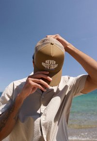 Homme sur la plage ajustant une casquette beige portant l'inscription « Ne jamais gaspiller le talent », vêtu d'une chemise légère texturée avec l'océan et un ciel dégagé en arrière-plan.