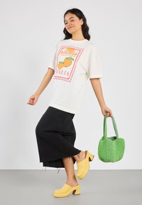 White t-shirt with orange fruit graphic, black denim skirt with frayed hem, yellow clogs, and green textured handbag.