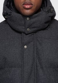 Mörkgrå ytterkläder med hög krage, utrustade med tryckknappar och dragsko. Materialet verkar texturerat och ullliknande med en strukturerad design.