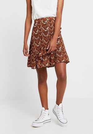 Kobieta ubrana w brązową spódnicę o wzorze wiązaną na zakładkę, biały bezrękawnik i białe wysokie trampki, stojąca na tle gładkiego tła.