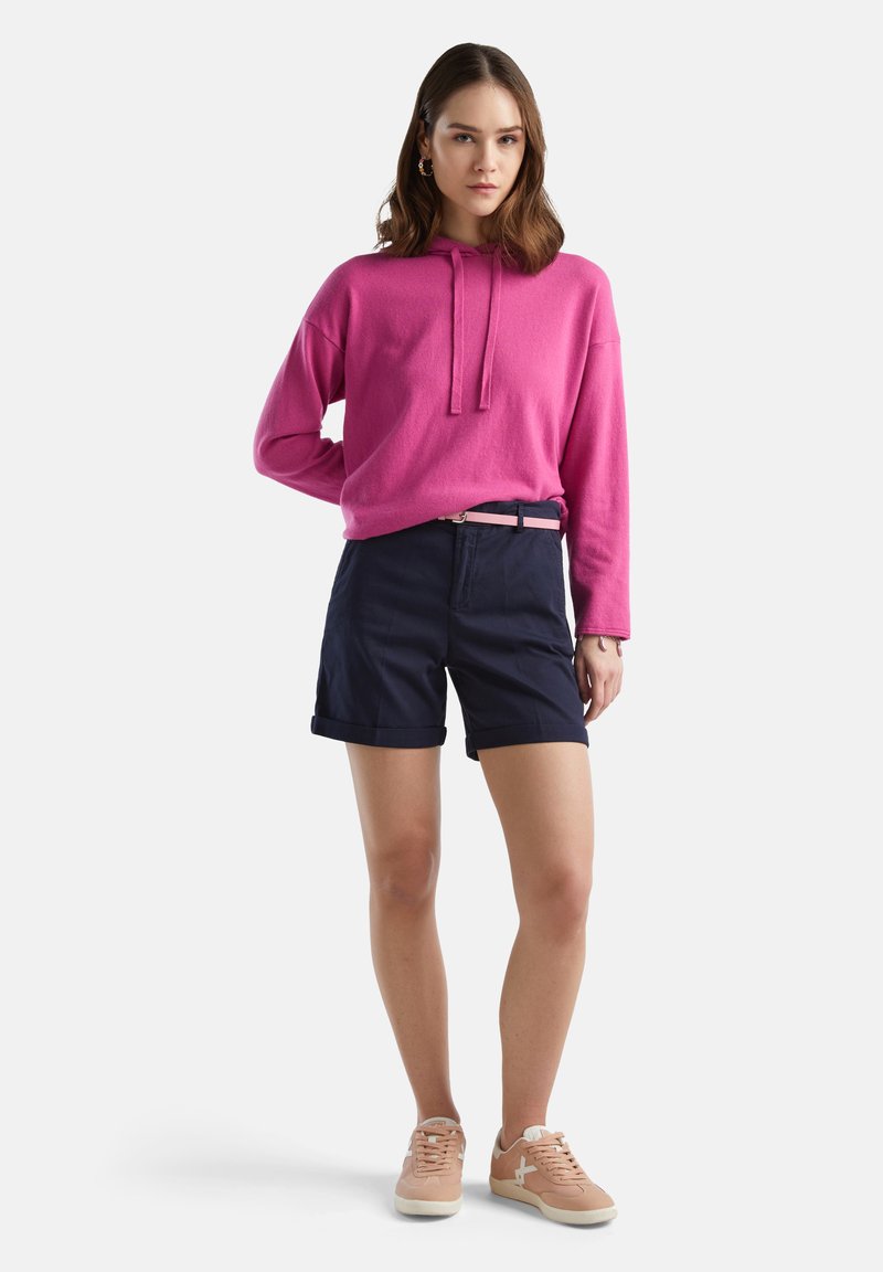 Różowa bluza z kapturem, granatowe szorty z podwiniętymi nogawkami i beżowe trampki z białymi akcentami. Swobodny zestaw w luźnym fasonie.