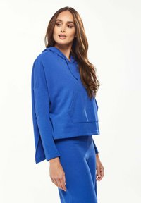 Blue hoodie made of soft fabric, featuring a front pocket, a relaxed fit, and long sleeves with rolled cuffs. Paired with matching blue bottoms.