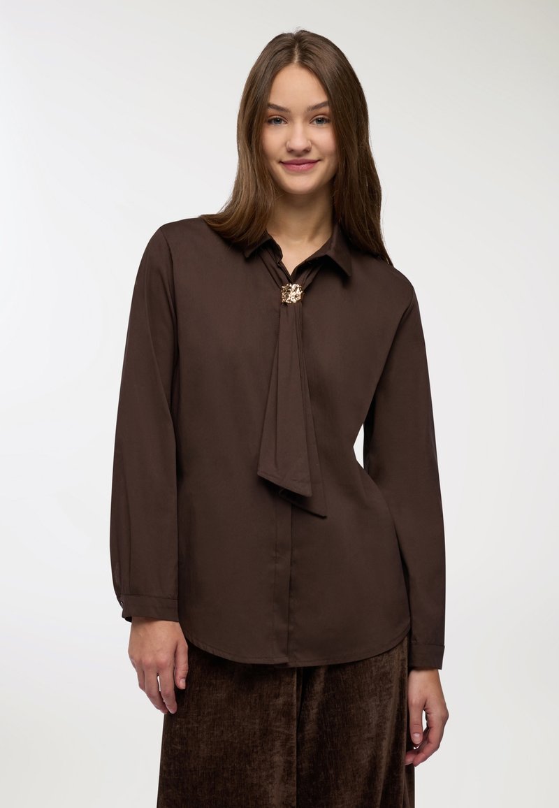 Giovane donna con lunghi capelli castani indossa una blusa marrone scuro con una spilla decorativa e pantaloni di velluto marrone larghi, sorridendo dolcemente.