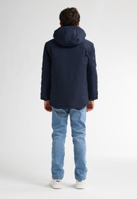 Giacca imbottita blu navy con cappuccio, polsini elasticizzati e una texture liscia. Abbinata a jeans azzurri e sneaker bianche.