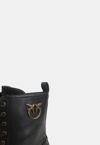 Parte superior de bota de cuero negro con detalle de anillo de metal dorado que presenta dos pájaros que se enfrentan uno al otro en el lateral cerca de los cordones.