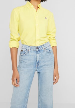 Persoon draagt een felgeel overhemd met opgerolde mouwen, ingestopt in een hooggesneden lichtblauwe spijkerbroek.