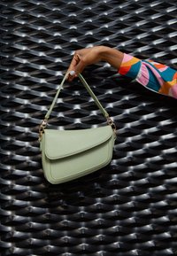 Mano con smalto per unghie blu pastello che tiene una borsa in pelle verde chiaro contro uno sfondo in metallo nero testurizzato.