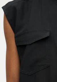 Λεπτομέρεια μαύρου ρούχου χωρίς μανίκια που δείχνει τσέπη με καπάκι και ορατές ραφές στην περιοχή του άνω βραχίονα σε ανοιχτό φόντο.