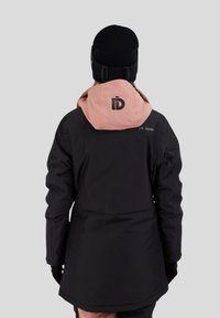 Sort vandtæt jakke med en lyserød hætte, med et logo på ryggen, lange ærmer og tætsiddende design. Båret med en sort beanie.