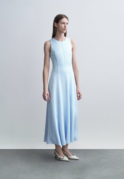 Femme debout portant une robe sans manches bleu clair jusqu'aux chevilles avec des talons blancs à bouts pointus, sur un fond uni clair.