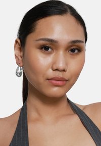 Boucles d'oreilles en argent en forme de goutte, avec une surface polie, reflétant la lumière, portées par un mannequin aux longs cheveux noirs et portant un haut à col haut à rayures.
