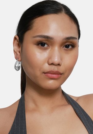 Orecchini a forma di goccia d'argento con superficie lucida che riflette la luce, indossati da un modello con lunghi capelli scuri e un top a righe sottili con colletto alto.