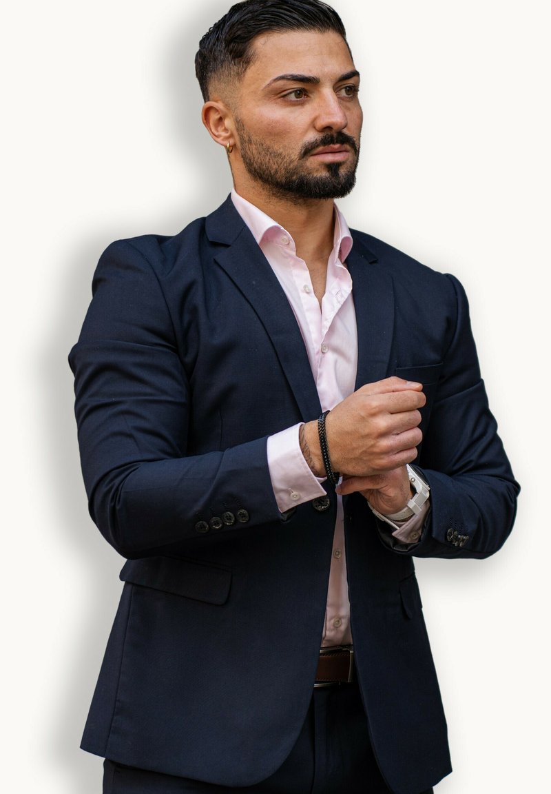 Navy blue suit jacket over a light pink shirt, with silver buttons. Black braided bracelet on wrist and white watch visible. Hands clasped.
