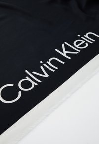 Must musträt, millel keskosas on valge logo "Calvin Klein", valge serva ja kulunud ääred. Sile tekstuur.