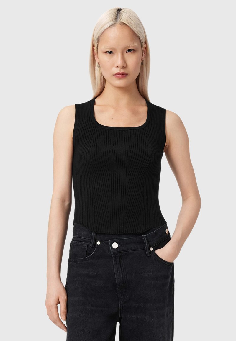Femme aux cheveux blonds raides portant un haut noir côtelé sans manches et un jean noir taille haute, une main dans la poche.