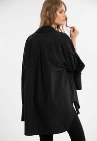Sort oversized skjorte med krave, lange ærmer og siderifts. Den glatte tekstur og afslappede pasform fremhæver dens moderne design.