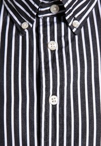 Czarna i biała koszula z guzikami, z wywiniętym kołnierzykiem i białymi plastikowymi guzikami z przodu, charakteryzująca się gładką teksturą materiału.