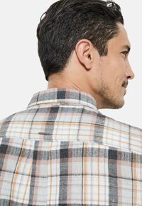 Chemise à carreaux présentant un mélange de couleurs blanc, gris, noir et orange. Les détails remarquables comprennent un petit logo au dos et un col classique.