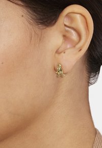 El pendiente de dinosaurio en tono dorado presenta un diseño detallado, con un acabado pulido, que se adhiere al lóbulo de la oreja, mostrando una forma de animal caprichosa.