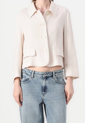 Donna che indossa una giacca beige corta con bottoni e tasche con pattina e jeans azzurri a vita alta su sfondo bianco.