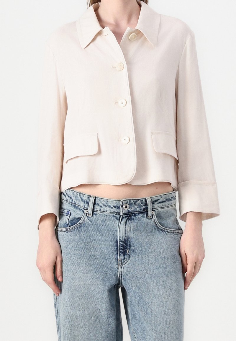 Femme portant une veste beige courte boutonnée avec des poches à rabat et un jean taille haute bleu clair sur fond blanc.