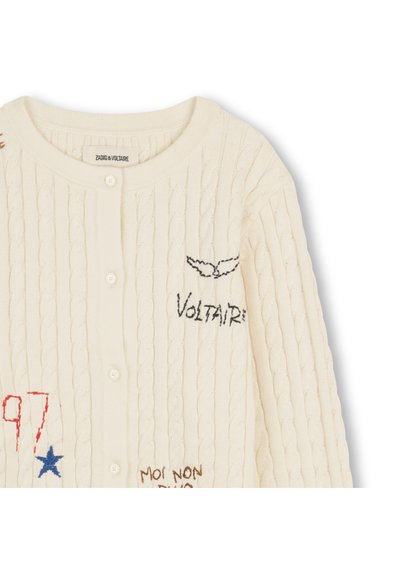 Ζακέτα πλεκτή σε κρεμ απόχρωση με σχέδιο με πλεξούδες, διακοσμημένη με κεντητό κείμενο και γραφικά σε μπλε, κόκκινο και μαύρο. Κλείσιμο με κουμπιά μπροστά.