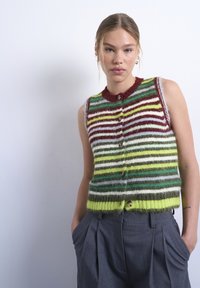 Νεαρή γυναίκα με ανοιχτά μαλλιά φοράει πολύχρωμο ριγωτό αμάνικο χνουδωτό γιλέκο με κουμπιά και γκρι πλισέ παντελόνια, με τα χέρια στ ις τσέπες.