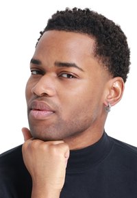 Orecchino a cerchio argento con accenti di pietre preziose trasparenti, indossato con un top nero a collo alto; capelli corti e strutturati di colore scuro; sfondo neutro.