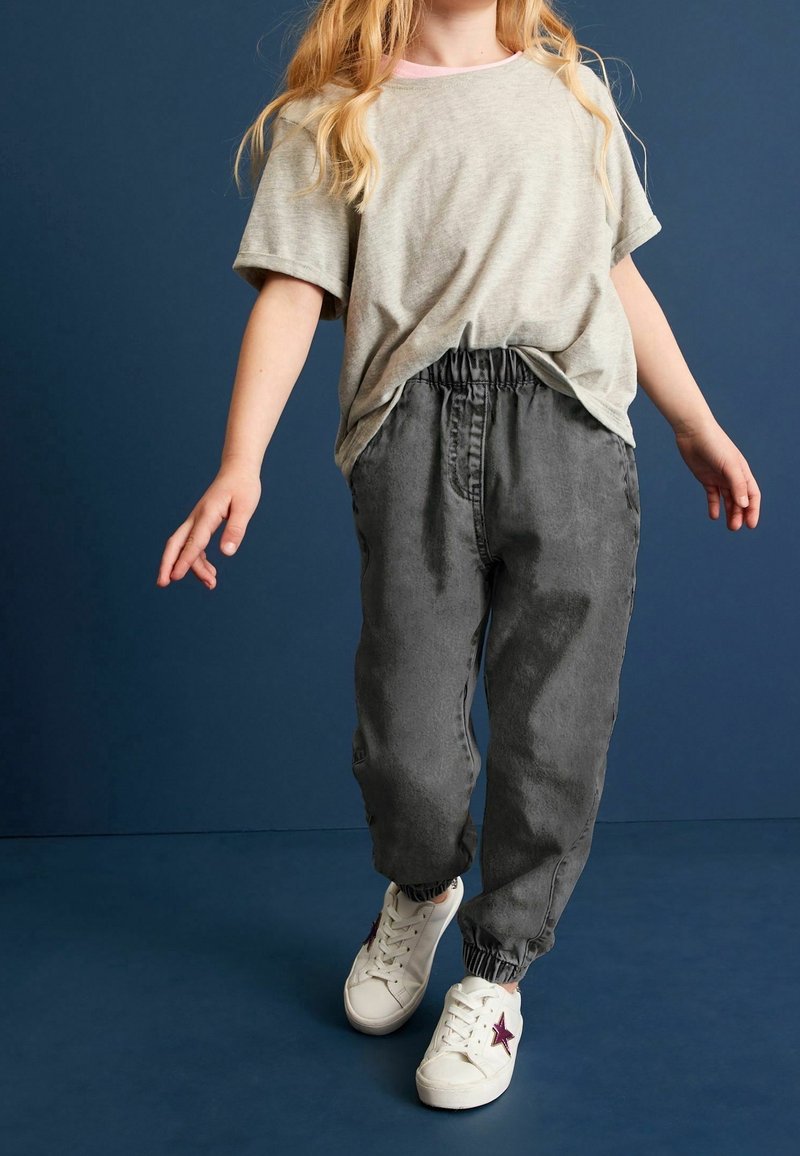 Enfant portant un t-shirt gris surdimensionné, un pantalon de jogging gris foncé et des baskets blanches avec une étoile violette sur fond bleu foncé.