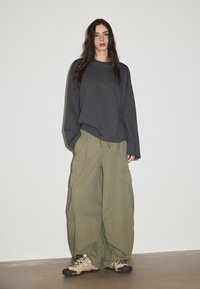 Woman with long dark hair wears a loose dark gray long-sleeve shirt, wide olive cargo pants, and beige hiking shoes, standing against a plain wall.