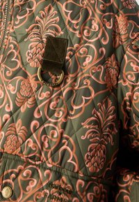 Casaco acolchoado em verde oliva com padrões florais em rosa e de abacaxi. Apresenta um laço de veludo e um destaque de ferragem da marca.