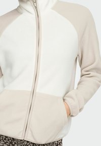 Chaqueta de forro polar con cremallera en color crema y beige con mangas ranglán, bolsillos tipo canguro y textura suave, adecuada para uso casual.