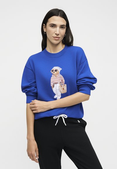 Νέα γυναίκα φοράει μπλε φούτερ με εκτύπωση σκύλου με γυαλιά ηλίου και καθημερινά ρούχα στο εμπρός μέρος, σε συνδυασμό με μαύρο παντελόνι.